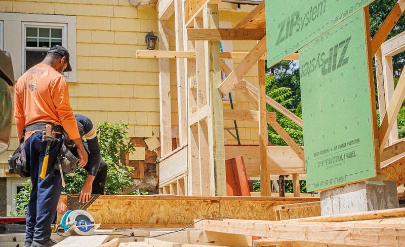 Construction workers building a wood-framed home addition next to a yellow house located in the Greater Boston area