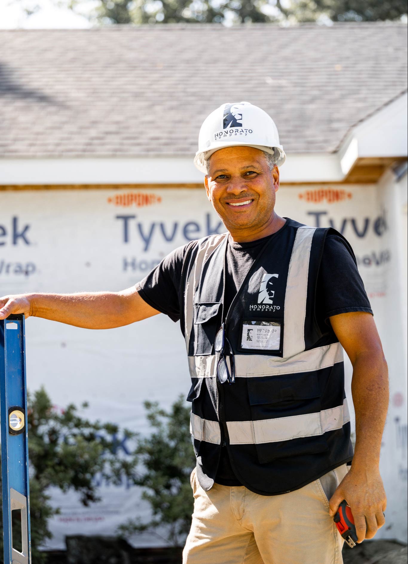 Smiling Honorato Company construction worker in safety gear standing at job site with house under renovation