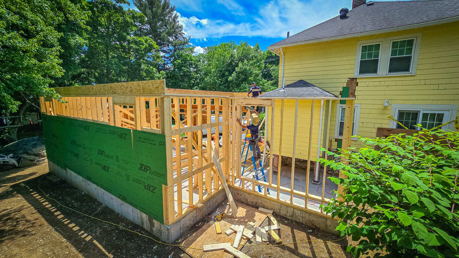 Greater Boston home addition under construction with wood framing, workers on ladders, and existing yellow house beside the new structure