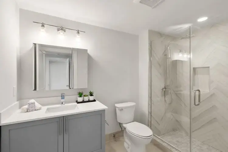 Bright modern bathroom with gray vanity, mirror lighting, toilet, and glass walk-in shower with marble tile in a home in Greater Boston