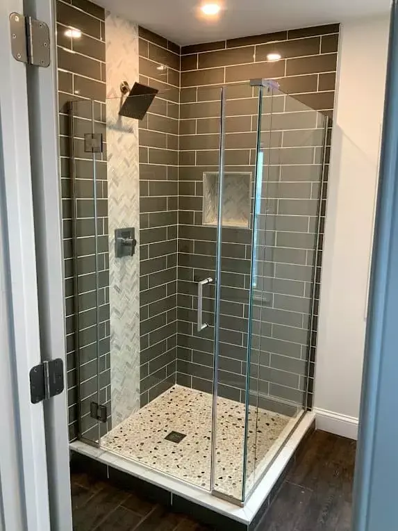 Modern glass walk-in shower with dark subway tile walls, rainfall showerhead, and pebble-style floor tile in a renovated bathroom in Greater Boston