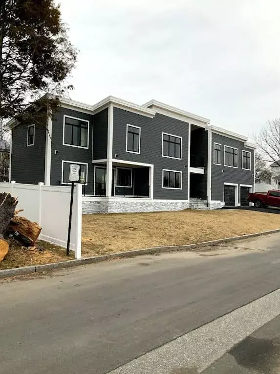 Modern multi-unit residential building with gray siding, large windows, and stone base along a quiet street in Greater Boston