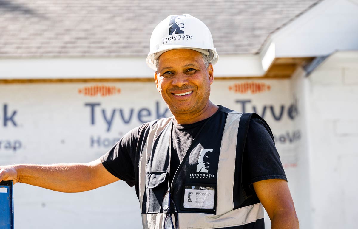 Smiling Honorato Company construction worker in safety gear standing at job site with house under renovation-1 Smiling Honorato Company construction worker in safety gear standing at job site with house under renovation-1